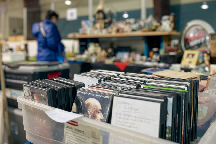 Secondhand items on display at the T.G. Smith Arena during the Steinbach Community Garage Sale with a person browsing additional items in the background.